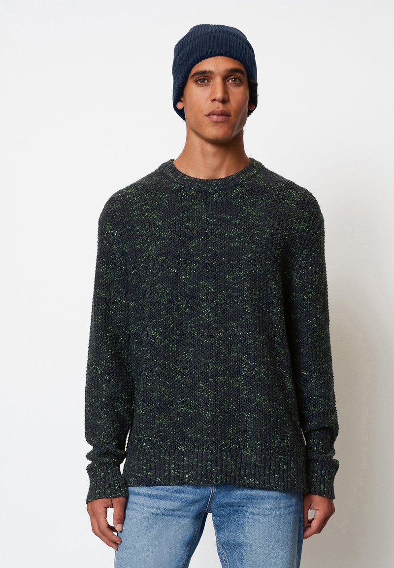Pull en tricot texturé vert foncé et bleu marine avec col rond et manches longues, associé à un jean bleu clair.