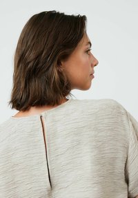 Haut texturé beige clair avec un col rond et un détail en œillet au dos. Manches courtes et coupe décontractée.