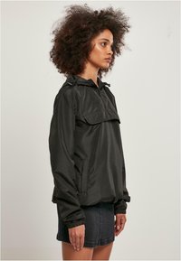 Zwarte pulloverjack met capuchon, voorzien van een voorzak en elastische manchetten. Glad textuur en lichtgewicht materiaal. Gestyled met een denimrok.