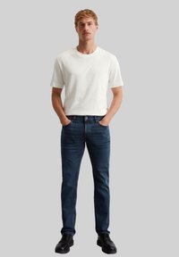 Witte korte mouwen t-shirt gecombineerd met donkerblauwe jeans. Het model heeft de handen in de zakken en draagt zwarte veterschoenen. Simpele, casual stijl.