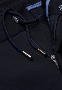 Gros plan sur un sweat à capuche marine, présentant un tissu texturé, des cordons avec embouts métalliques et une poche zippée.