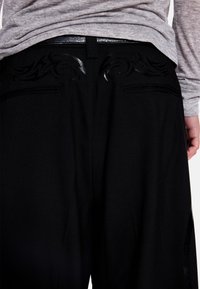 Juodos kelnės su glotniu audiniu, nugaroje puošnios blizgios detalės ir dvi galinės kišenės. Juosmuo papuoštas tamsia diržu.