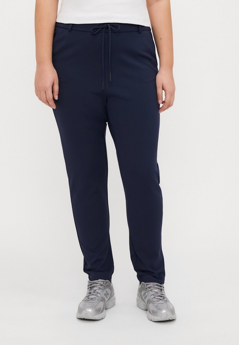 Personne portant un pantalon à cordon de serrage bleu marine, un haut blanc et des chaussures de sport argentées, debout devant un fond uni.