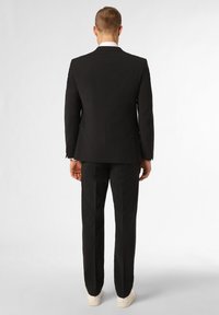 Schwarzer Anzug mit passender Hose, taillierte Passform, einreihiges Design und weißes Hemd. Sneaker sichtbar, glatte Stofftextur.