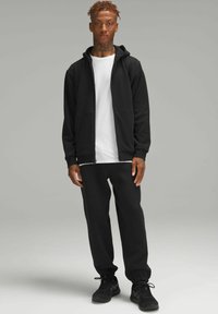 Felpa nera con zip e due tasche frontali, abbinata a pantaloni jogger neri. Modello che indossa una maglietta bianca sotto e scarpe sportive nere.