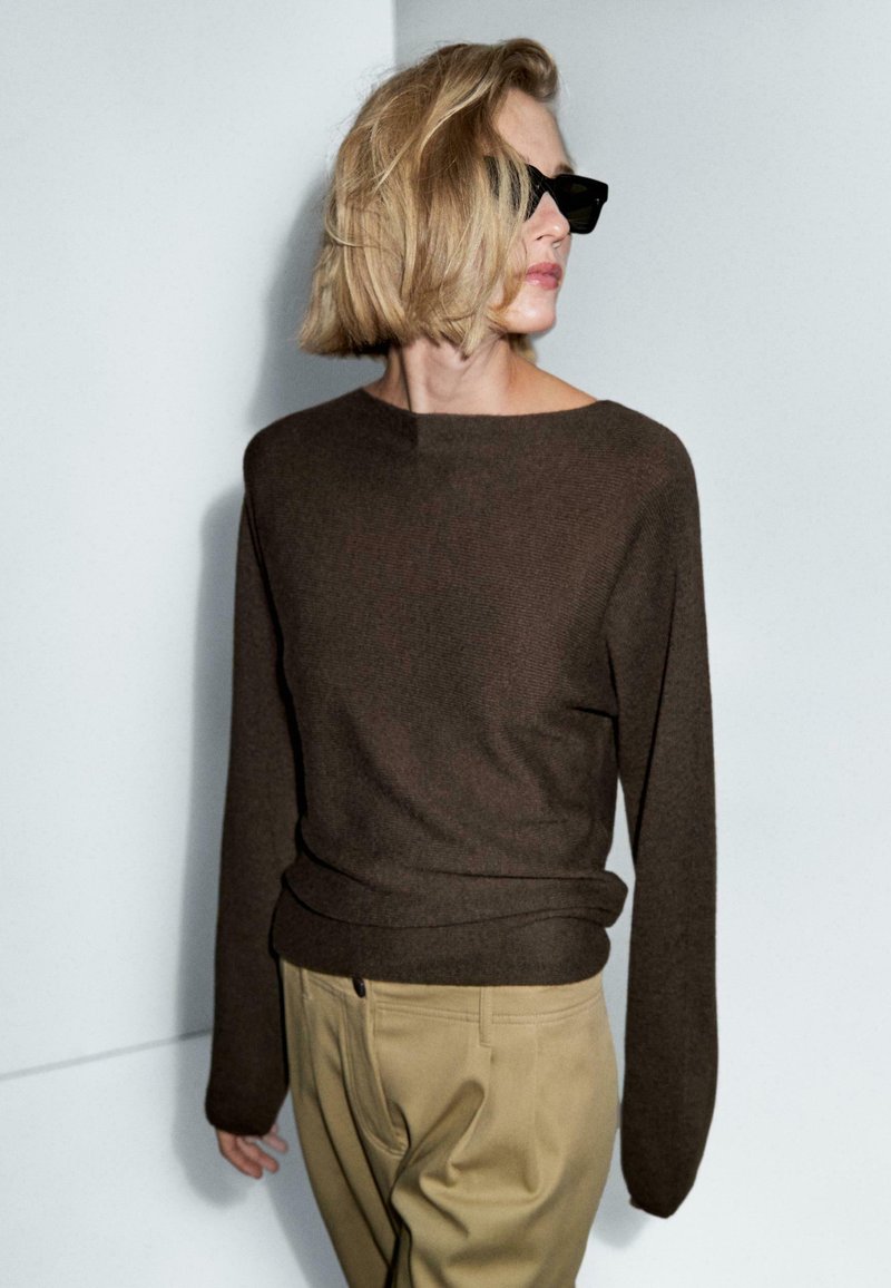 Persoon met kort blond haar, die zwarte zonnebril, een donkerbruine lange mouwen trui en beige broek draagt, terwijl hij naar de zijkant kijkt tegen een effen muur.