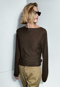 Persona con capelli biondi corti, che indossa occhiali da sole neri, un maglione a maniche lunghe marrone scuro e pantaloni beige, che guarda di lato contro un muro neutro.