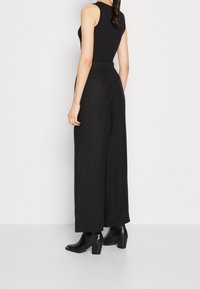 Černý top bez rukávů spárovaný s širokými černými kalhotami. Kalhoty mají vysoký pas a hladkou texturu, doplněné o černé boty na podpatku.