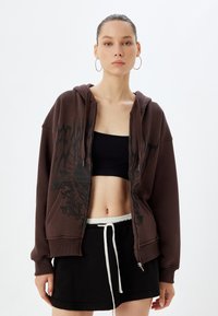Felpa marrone con zip e design grafico in nero, presenta una vestibilità ampia, grandi tasche e un orlo con cordoncino. Abbinate a un top corto nero e una gonna.