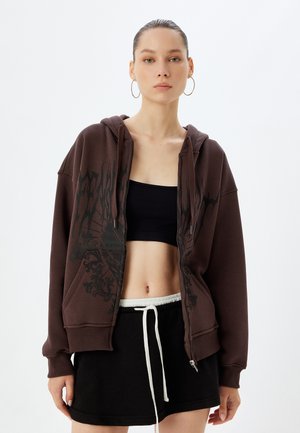 Brun hættetrøje med lynlås og en grafisk design i sort. Den har en løs pasform, store lommer og en snøre i kanten. Stylet med en sort crop top og nederdel.