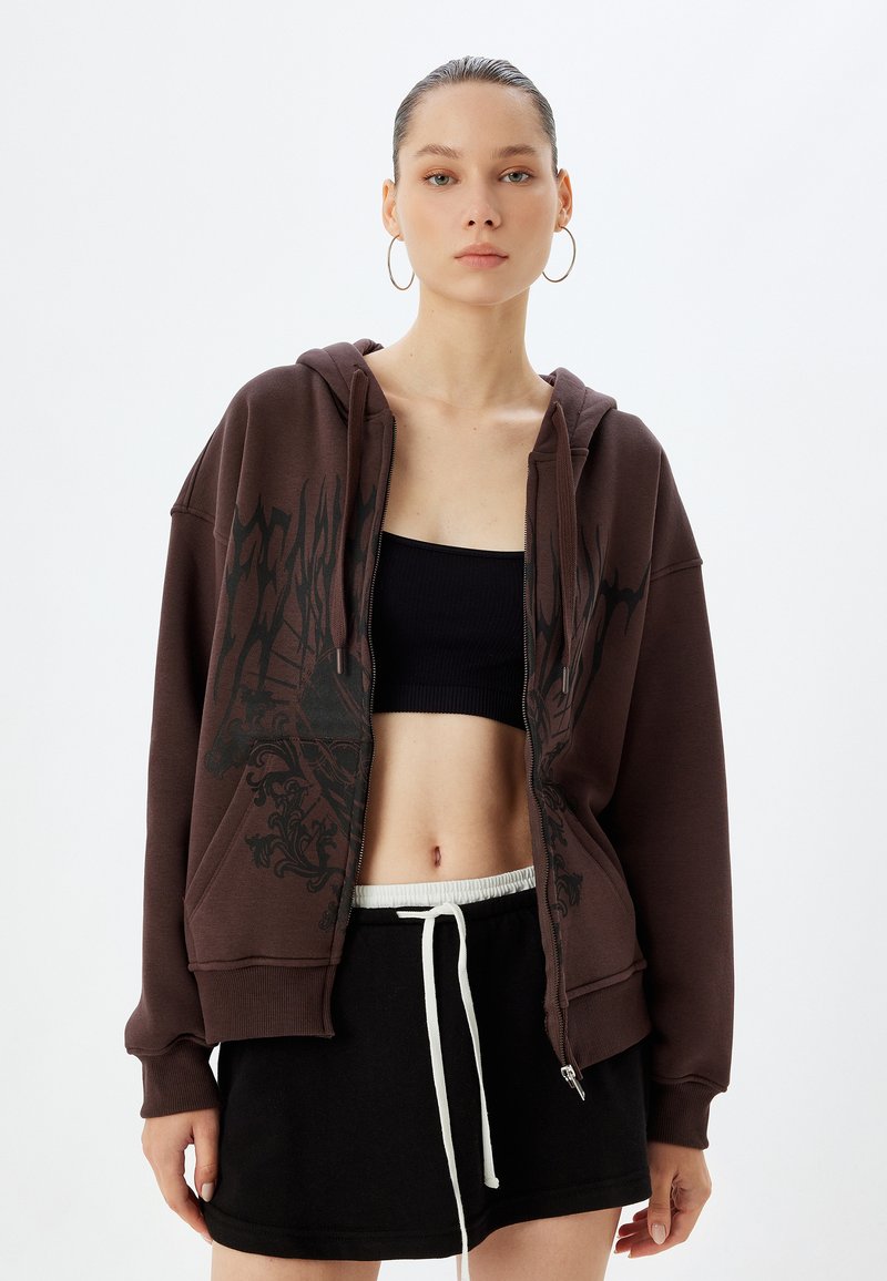 Felpa marrone con zip e design grafico in nero, presenta una vestibilità ampia, grandi tasche e un orlo con cordoncino. Abbinate a un top corto nero e una gonna.