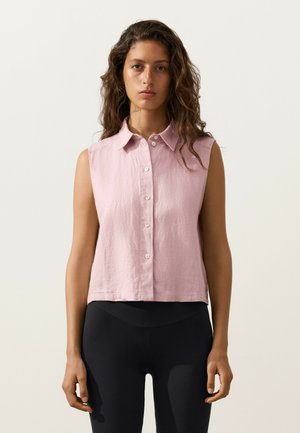 Femme aux longs cheveux bouclés portant une chemise sans manches rose clair boutonnée et un pantalon noir ajusté sur un fond uni.
