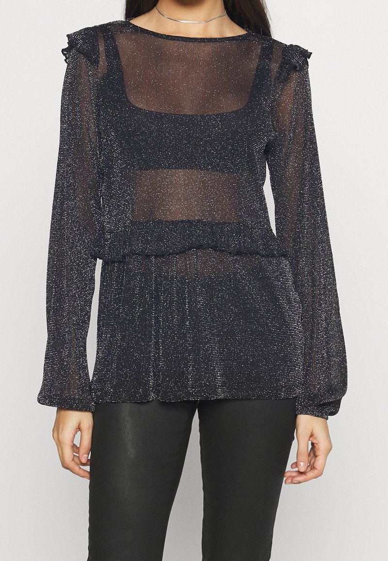 Blusa nera trasparente a maniche lunghe con riflessi metallici, spalle arruffate e vestibilità ampia, indossata sopra un top semplice, abbinata a pantaloni neri.
