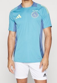 Vaaleansininen urheilupaita lyhyillä hihoilla, jossa on Adidas-logo, Ajaxin tunnus sekä kontrastoivia tummansinisiä yksityiskohtia, valmistettu hengittävästä kankaasta.