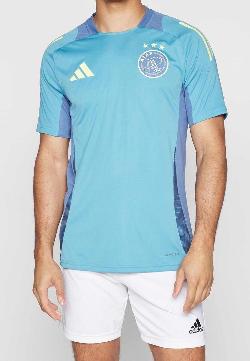 Vaaleansininen urheilupaita lyhyillä hihoilla, jossa on Adidas-logo, Ajaxin tunnus sekä kontrastoivia tummansinisiä yksityiskohtia, valmistettu hengittävästä kankaasta.