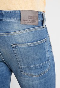 Vaqueros de mezclilla en azul claro, con un bolsillo trasero y una etiqueta de cuero gris texturizado con "Scotch & Soda" grabado en ella.