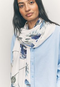 Écharpe fleurie blanche avec des motifs bleus et verts, drapée autour du cou, associée à une chemise à boutons bleu clair. Texture de tissu douce.