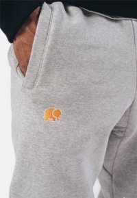 Jogging en coton gris avec poches latérales, arborant un logo d'éléphant brodé orange sur la cuisse. Texture douce et coupe décontractée.