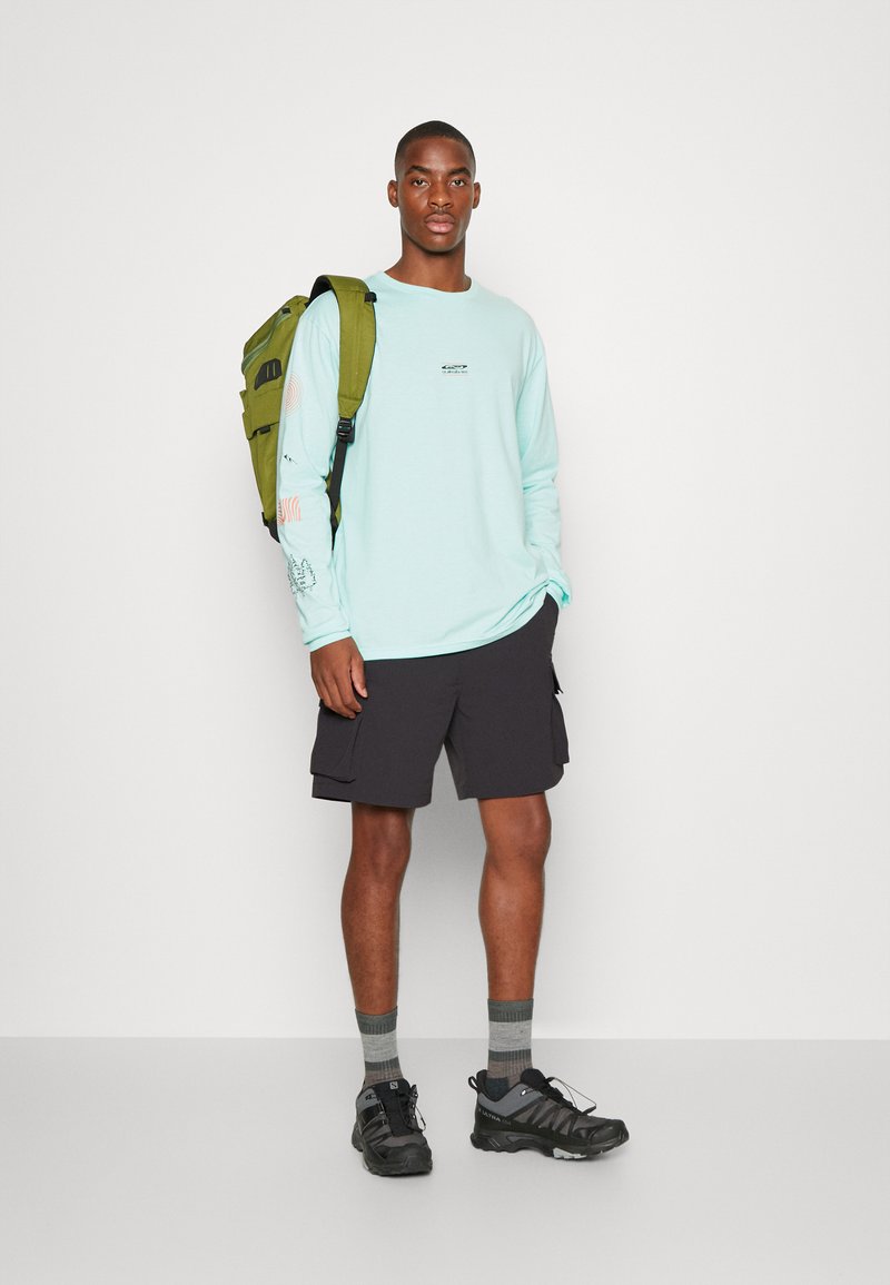 Light blue long-sleeve shirt with graphic details, black cargo shorts, gray socks, and black hiking shoes; carrying a green backpack.