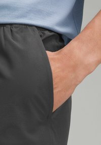 Sorte atletiske shorts med en glat tekstur, der har en sidelomme, elastisk talje og metalliske syninger. Lys blå skjorte ovenfor.