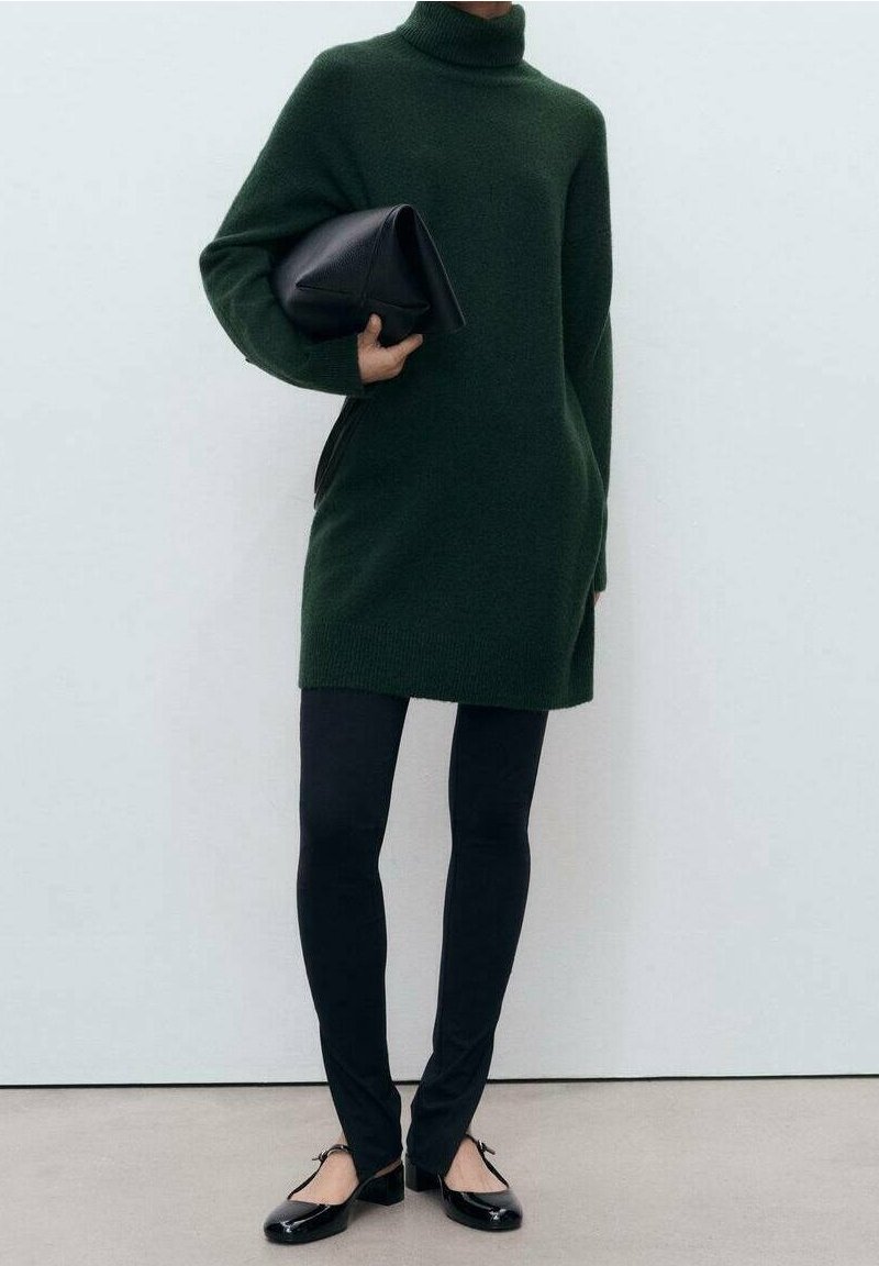 Personne portant une robe-pull à col roulé oversize vert foncé, des leggings noirs, des ballerines noires, et tenant une pochette noire.