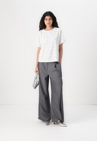 Blouse en eyelet blanc à manches courtes associée à un pantalon large gris. Le mannequin tient un sac à main plissé argenté et porte des chaussures métalliques.