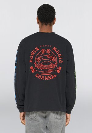 Person iført en sort langærmet skjorte med rød grafik af en frø og teksten "Edwin Music Channel" på ryggen, kombineret med grå jeans.