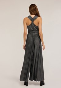Tuta metallizzata grigio scuro con collo halter annodato, gambe larghe plissettate e vita aderente, che mette in mostra una texture scintillante.