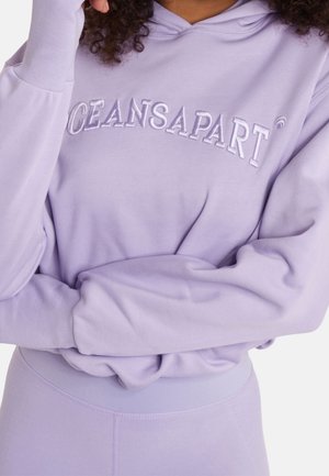 Person trägt einen lavendelfarbenen Hoodie mit "OCEANS APART" auf der Brust gestickt und passende lavendelfarbene Leggings, die Arme verschränkt.