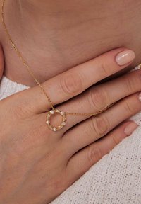 Złoty naszyjnik z okrągłym wisiorkiem ozdobionym małymi białymi perłami, noszony na odkrytej szyi, a ręka spoczywa na fakturowanym jasnym swetrze.