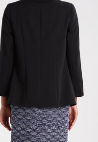 Frau in schwarzem, tailliertem Blazer und strukturiertem dunkelblauen und weißen Rock, von hinten gezeigt, steht vor einem einfarbigen Hintergrund.