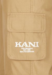 Svetlohnedé cargo šortky s prednou vreckom na nášivku a bielym vyšitým logom "KANI" a textom "THE ORIGINAL EST. 1989 REG. TRADEMARK" pod ním.