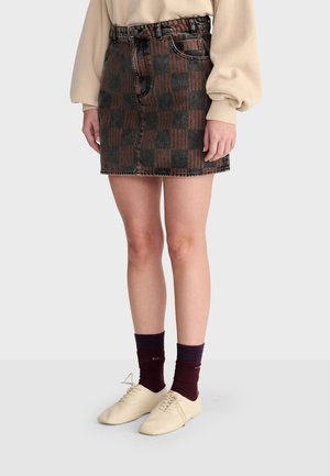 Personne portant une mini-jupe à carreaux marron et grise, un haut beige à manches longues, des chaussettes bordeaux et des chaussures crème à lacets, debout.