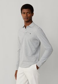 Muž v svetlosivej dlhorukávovej polo košeli s malým čiernym logom, bielych nohaviciach, stojaci pred jednoduchým svetlým pozadím.