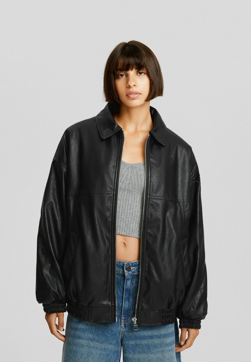 Chaqueta de cuero sintético negra con un ajuste relajado, cierre de cremallera en el frente y puños elásticos. Combinada con un top corto gris y jeans de tiro alto.