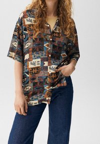 Chemise à manches courtes à boutons avec un motif géométrique multicolore dans des tons chauds ; le tissu semble texturé et léger. Portée avec un jean foncé.