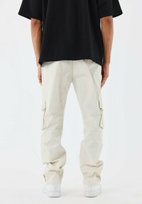 Pantalones cargo beige con dos bolsillos laterales, puños elásticos y una textura suave. Combinados con una camisa negra holgada y zapatillas blancas.