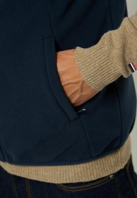 Marinblå fleecejacka med beige ribbade ärmslut och midja. Sidoficka och subtil detalj i metall nära fickan. Texturerad tygutseende.