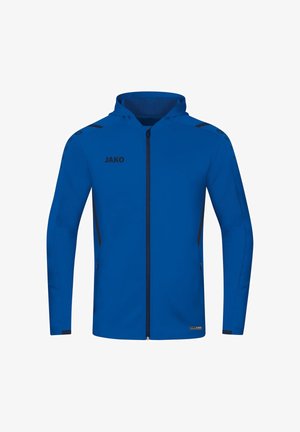 Mann trägt blauen Kapuzenpullover mit Reißverschluss und marineblaue Jogginghose, dazu schwarze Sneakers, steht und schaut nach vorne vor weißem Hintergrund.