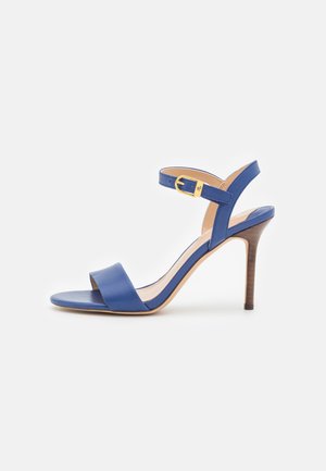 Blauwe sandaal met hoge hak, open teen, enkelbandje, gouden gesp en houten stilettohak op een witte achtergrond.