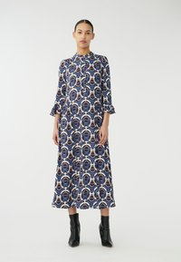 Långärmad klänning i marinblått med ett geometriskt mönster i beige och brunt. Har volang ärmar och hög krage, tillverkad av mjuk tyg.