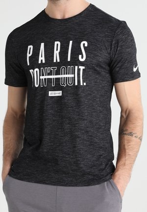 Mężczyzna w ciemnoszarej koszulce Nike z napisem "PARIS DON'T QUIT." i sloganem "JUST DO IT", ręce w kieszeniach szarych spodni.