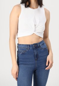 Top corto blanco sin mangas con un detalle de nudo en el frente, combinado con unos jeans de mezclilla azul de tiro alto que presentan un diseño clásico de cinco bolsillos.