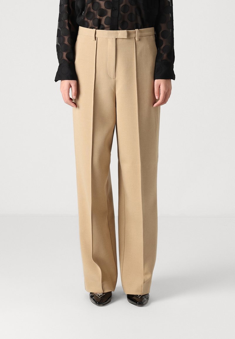 Pantalon beige à jambes larges porté avec une chemise noire transparente à boutons ornée de gros pois, associé à des chaussures noires pointues.