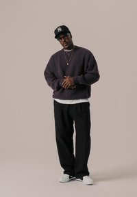 Dunkelvioletter Strickpullover, weißes T-Shirt darunter, schwarze weit geschnittene Hose, weiße Sneakers mit schwarzen Akzenten und eine schwarze Baseballkappe mit Logo.