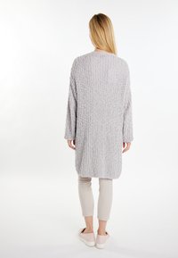 Cardigan lung tricotat de culoare gri deschis, cu un model texturat, croială lejeră, decolteu rotund și umeri căzuți, asortat cu pantaloni bej și teniși.
