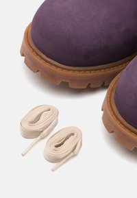 Fioletowe zamszowe buty na grubej, brązowej gumowej podeszwie. Dwa beżowe sznurowadła leżące pod butami, o płaskiej fakturze i zwężanych końcach.