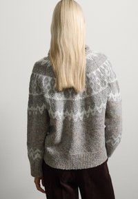 Pull en tricot gris avec un empiècement motif en blanc, comportant un ourlet en côte épais et un tissu texturé ; porté avec un pantalon en côtelettes marron.