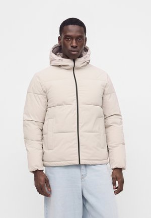 Homme portant une doudoune matelassée beige clair avec capuche et fermeture éclair frontale, associée à un jean bleu clair, debout devant un fond uni.