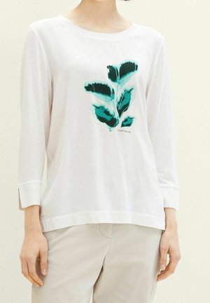 Camisa blanca de manga larga con estampado de hojas verdes en el pecho. Tejido suave y ligero con un corte relajado y aberturas laterales en el dobladillo.
